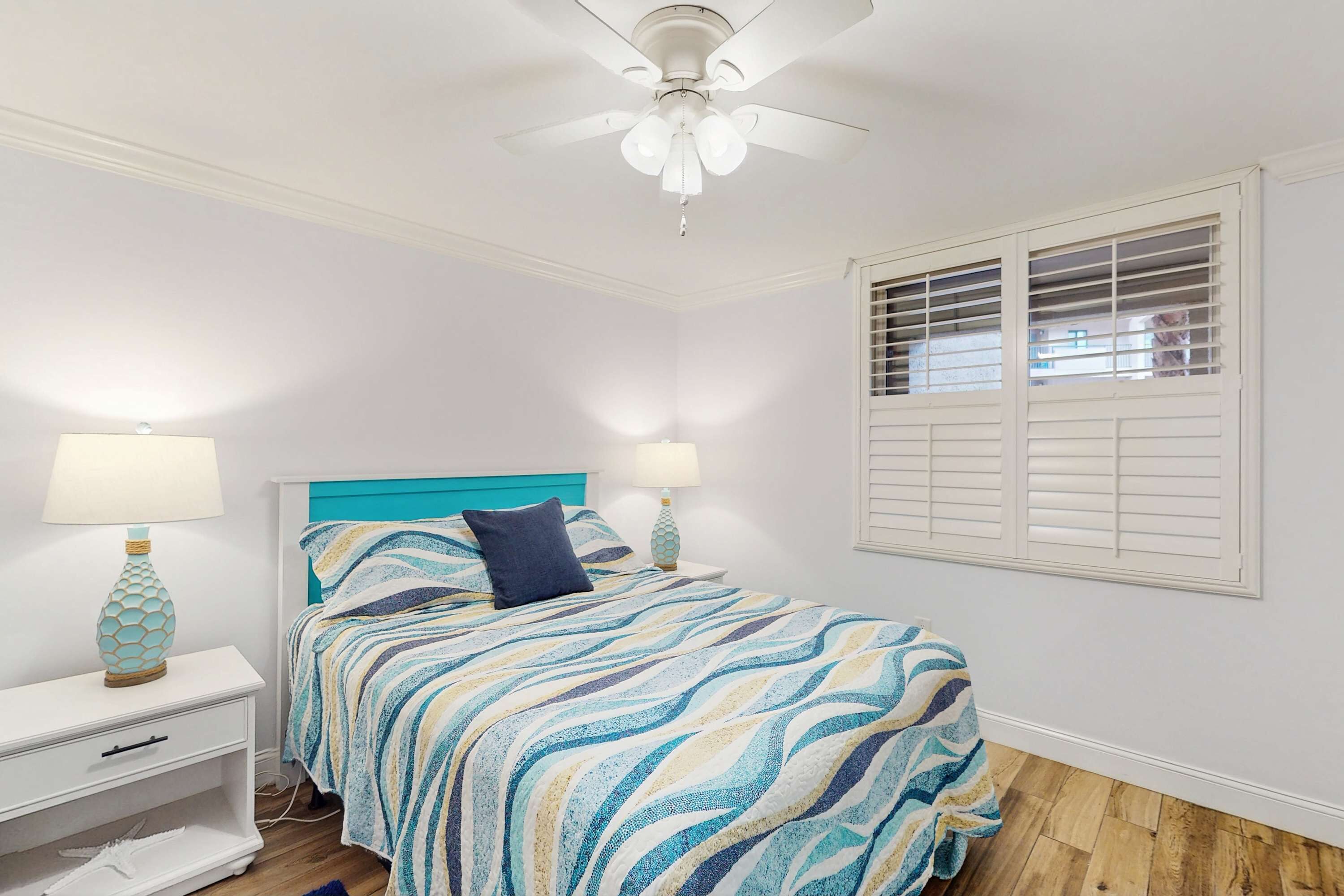 Une chambre à coucher avec un lit, des tables de chevet, un ventilateur de plafond et une fenêtre avec des stores.