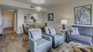 A living room with two sofas, a dining area with a table and chairs, a kitchen in the background, and a painting on the wall. - Beautiful condo with ocean & pool views, hot tub and snowbird friendly (Hilton Head)