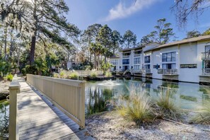 A wooden boardwalk leading to a building with multiple balconies overlooking a calm water body, surrounded by trees and vegetation. - Charming 1st floor condo near Coligny Beach Plaza and private beach access (Hilton Head)