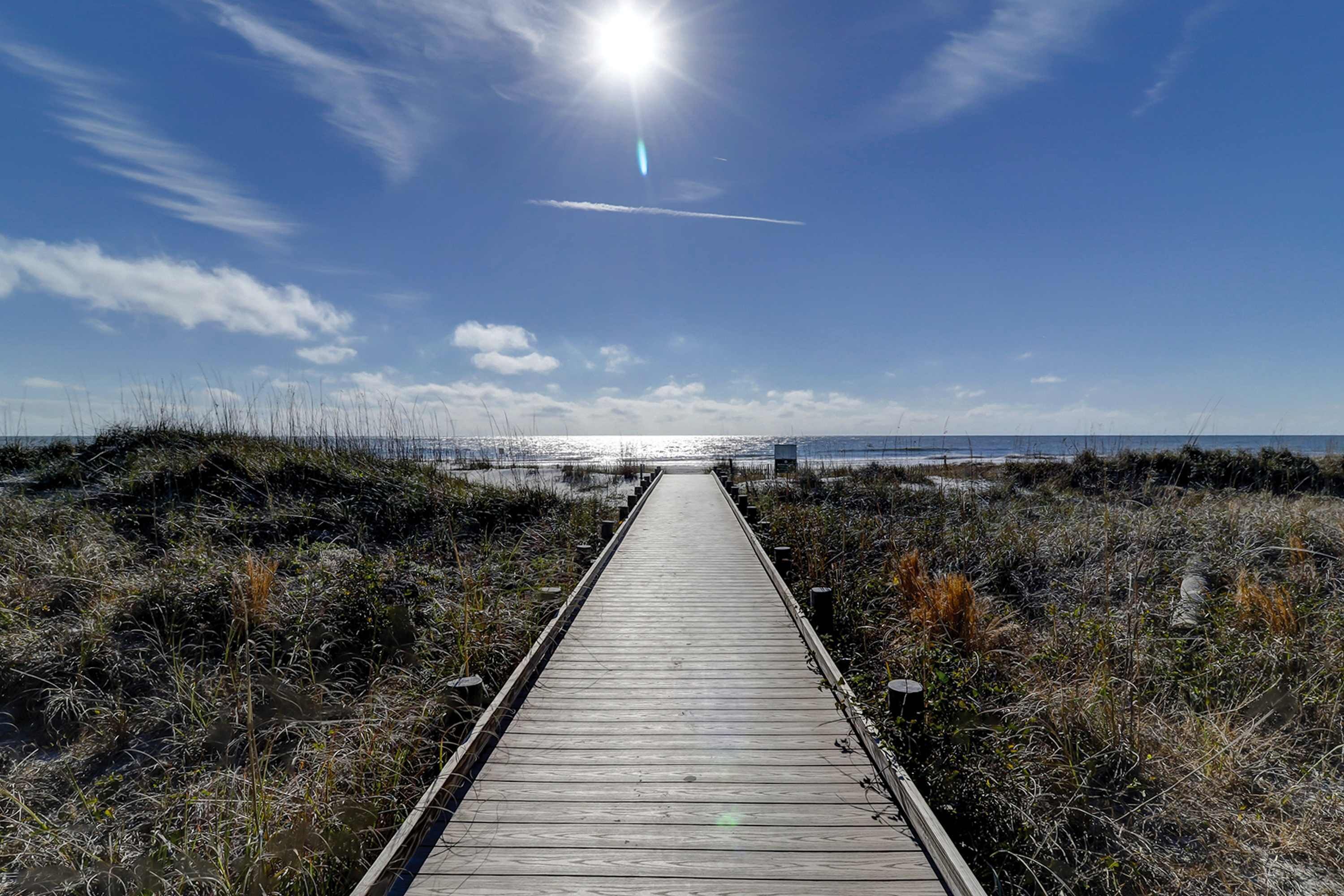 Ein hölzerner Steg führt zu einem Strand bei blauem Himmel und Sonnenschein.