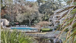 A pool with lounge chairs, a small pond, and a building with a balcony. - Spacious lagoon-view condo with tennis, outdoor pool, beach access, balcony (Hilton Head)