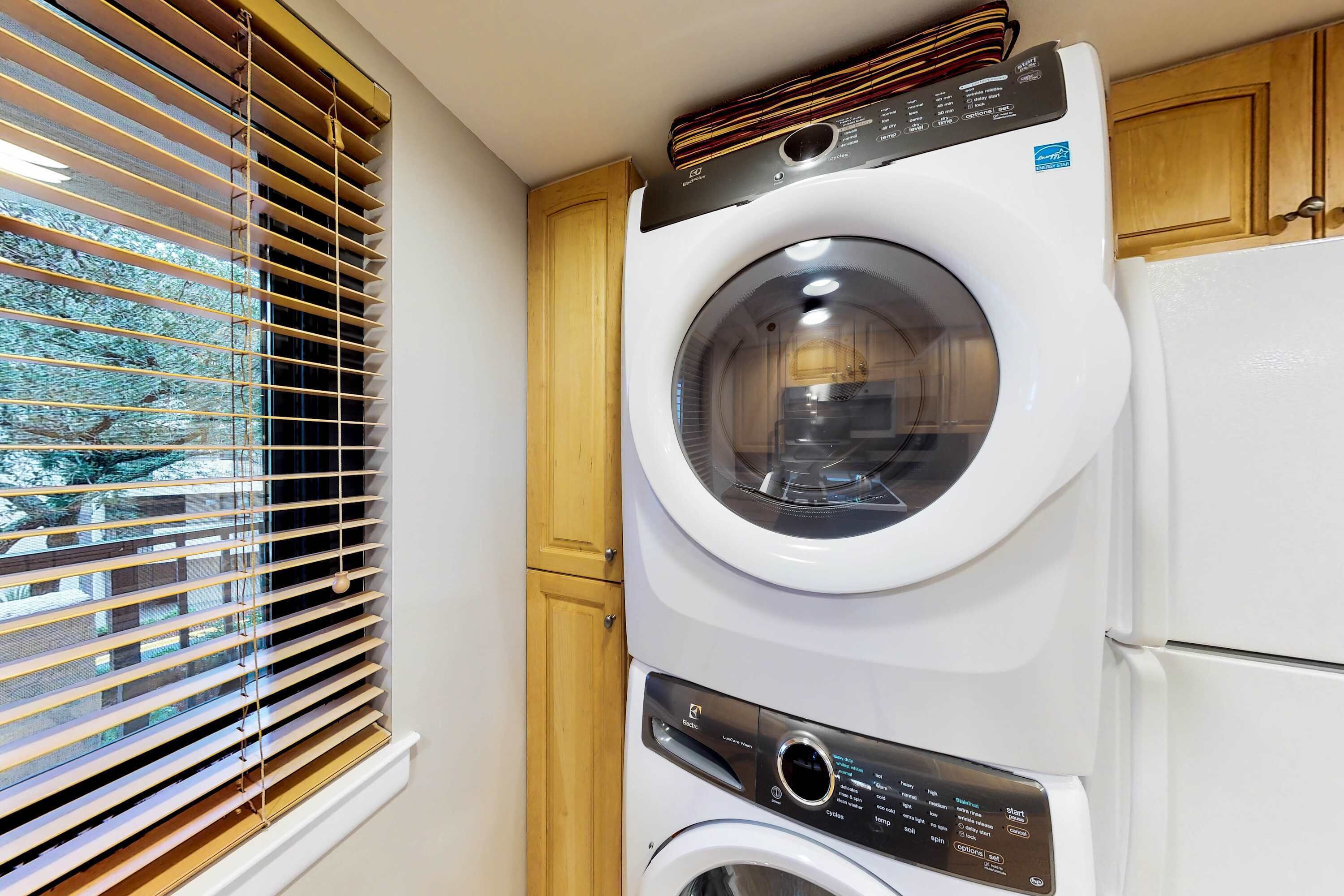 A compact laundry area with a front-loading washer and dryer, wooden cabinets, and a window with blinds.