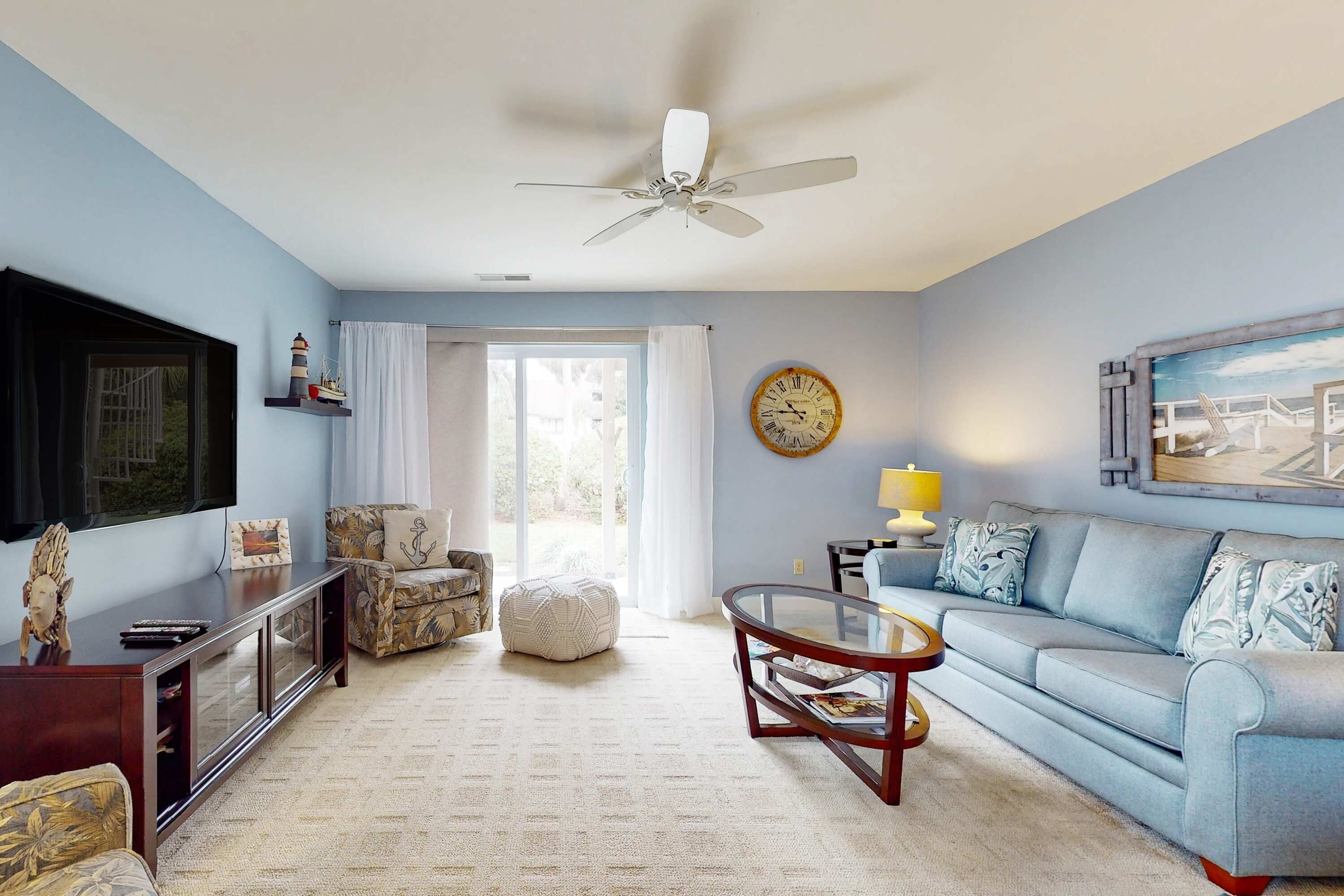 A living room with a sofa, a coffee table, a TV, a ceiling fan, a clock, and a window with curtains.