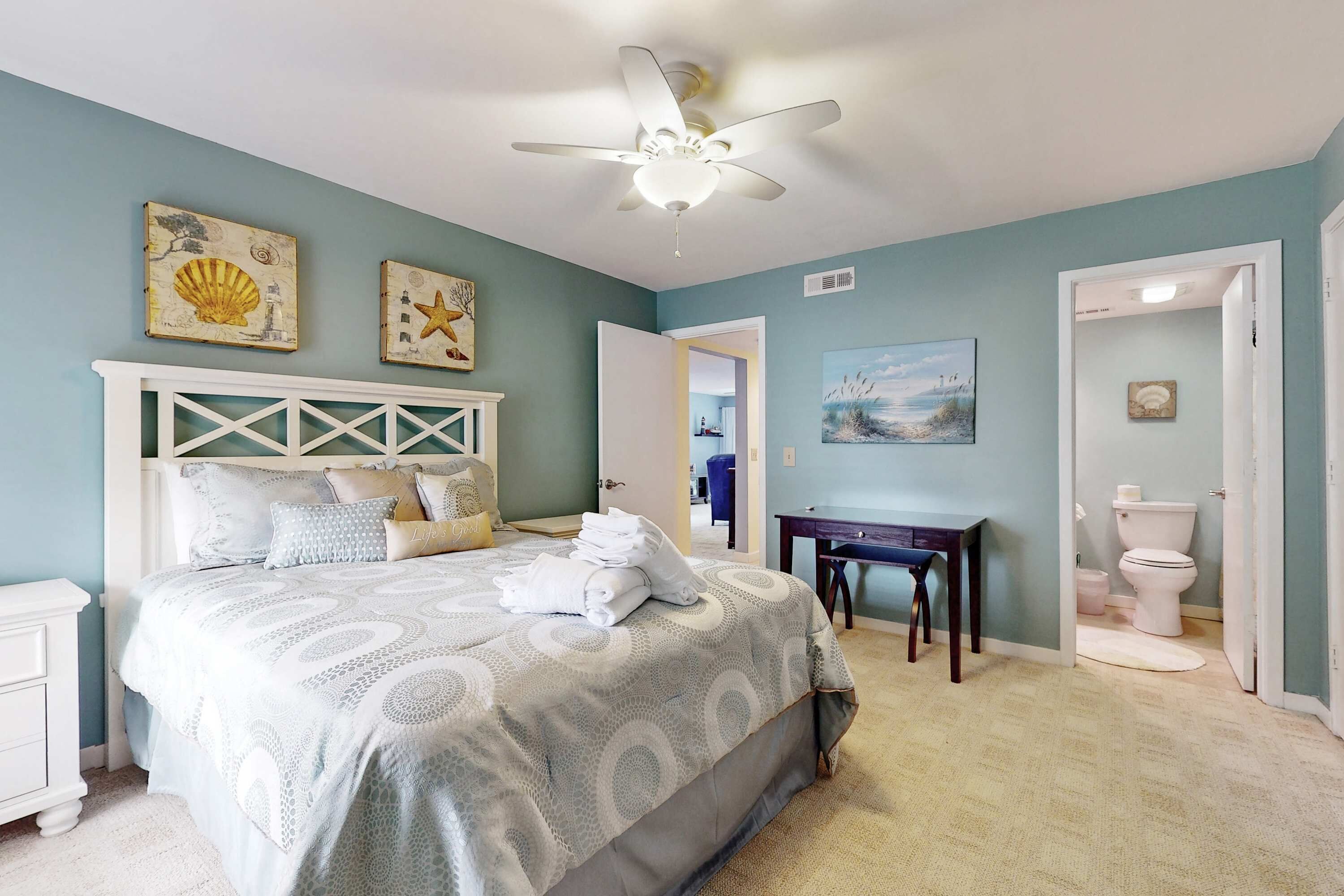 Une chambre à coucher avec un lit, un ventilateur de plafond, une table de chevet, une commode, un bureau et une salle de bain visible à travers une porte ouverte.