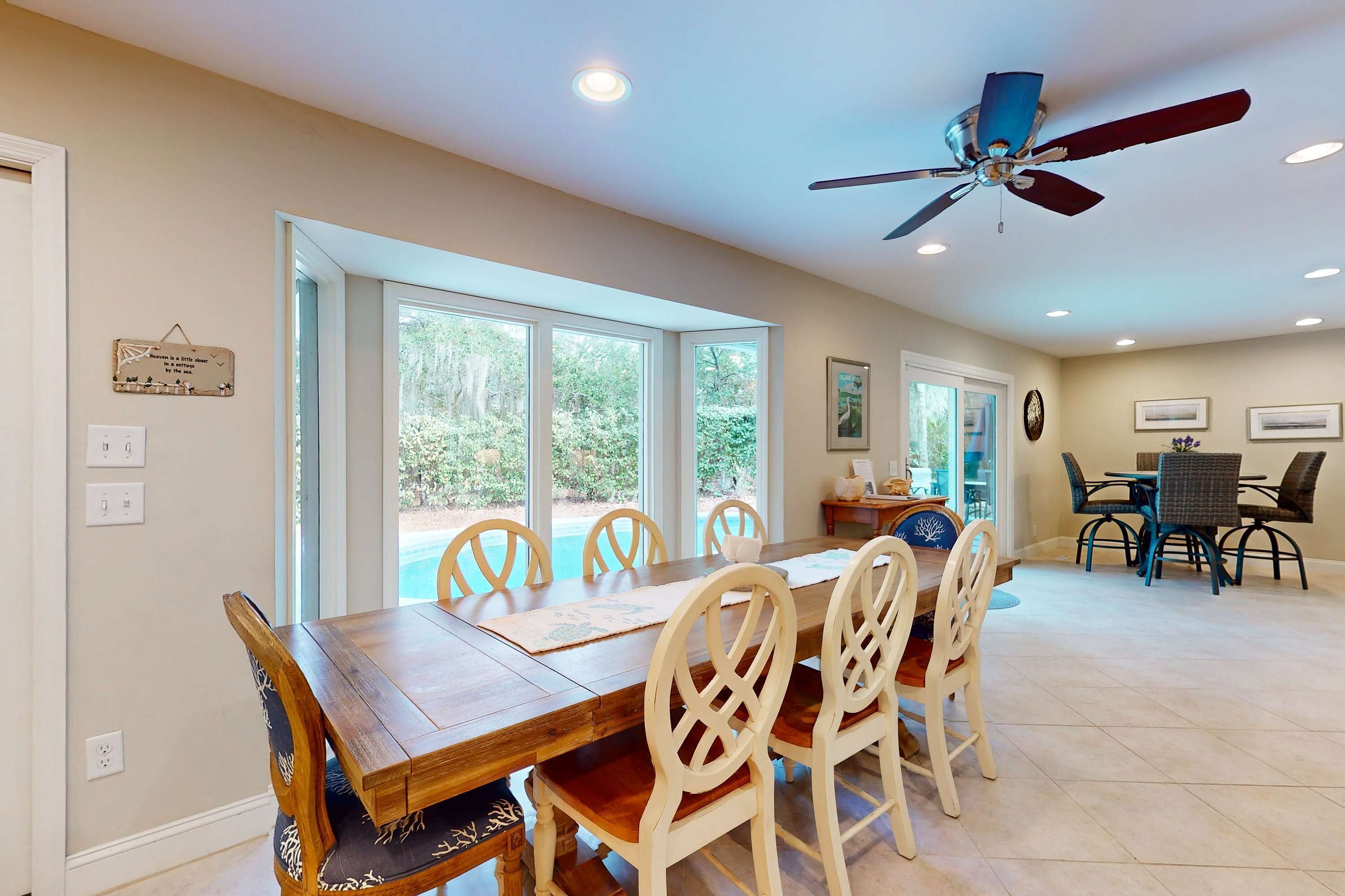 A dining area with a wooden table and chairs, large windows, a ceiling fan, and a view of trees.