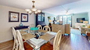 A dining area with a glass table, white chairs, a chandelier, framed artwork, and a view to the living room. - Updated Condo with Lagoon Views, Shared Pool, Hot Tub & WiFI - Near Beach (Hilton Head)
