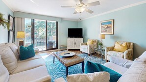 A living room with a ceiling fan, a television, a sofa, a chair, and a coffee table. - Updated Condo with Lagoon Views, Shared Pool, Hot Tub & WiFI - Near Beach (Hilton Head)