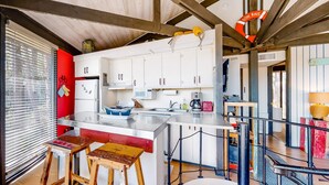 A modern kitchen with white cabinets, a stainless steel island, wooden stools, and a red accent wall. - Unique Treehouse Haven with Marsh-Front Views & Large Deck - Walk to Harbour Town (Hilton Head)