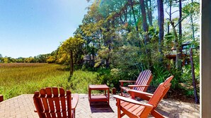 A patio with Adirondack chairs and a table, surrounded by a lush garden and a view of a field. - Unique Treehouse Haven with Marsh-Front Views & Large Deck - Walk to Harbour Town (Hilton Head)