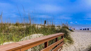 Una pasarela de madera que conduce a una playa con dunas de arena y vegetación escasa.