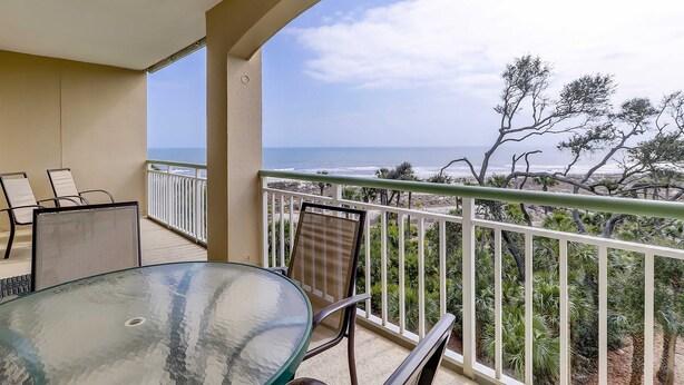 Un balcon avec une table et des chaises en verre, une balustrade blanche et une vue sur l’océan et les arbres.