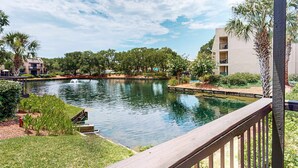 A serene outdoor pool area with a wooden deck, surrounded by lush greenery and a clear blue sky. - Roomy Villa with WiFi, Pool, & Hot Tub - Steps from the Ocean (Hilton Head)