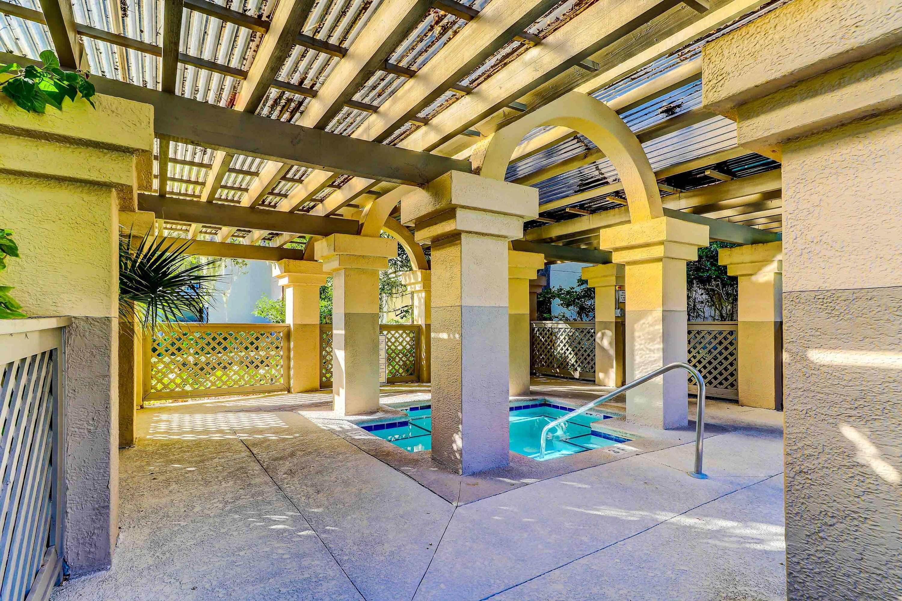 A covered outdoor swimming pool with a metal ladder, surrounded by columns and a wooden ceiling.