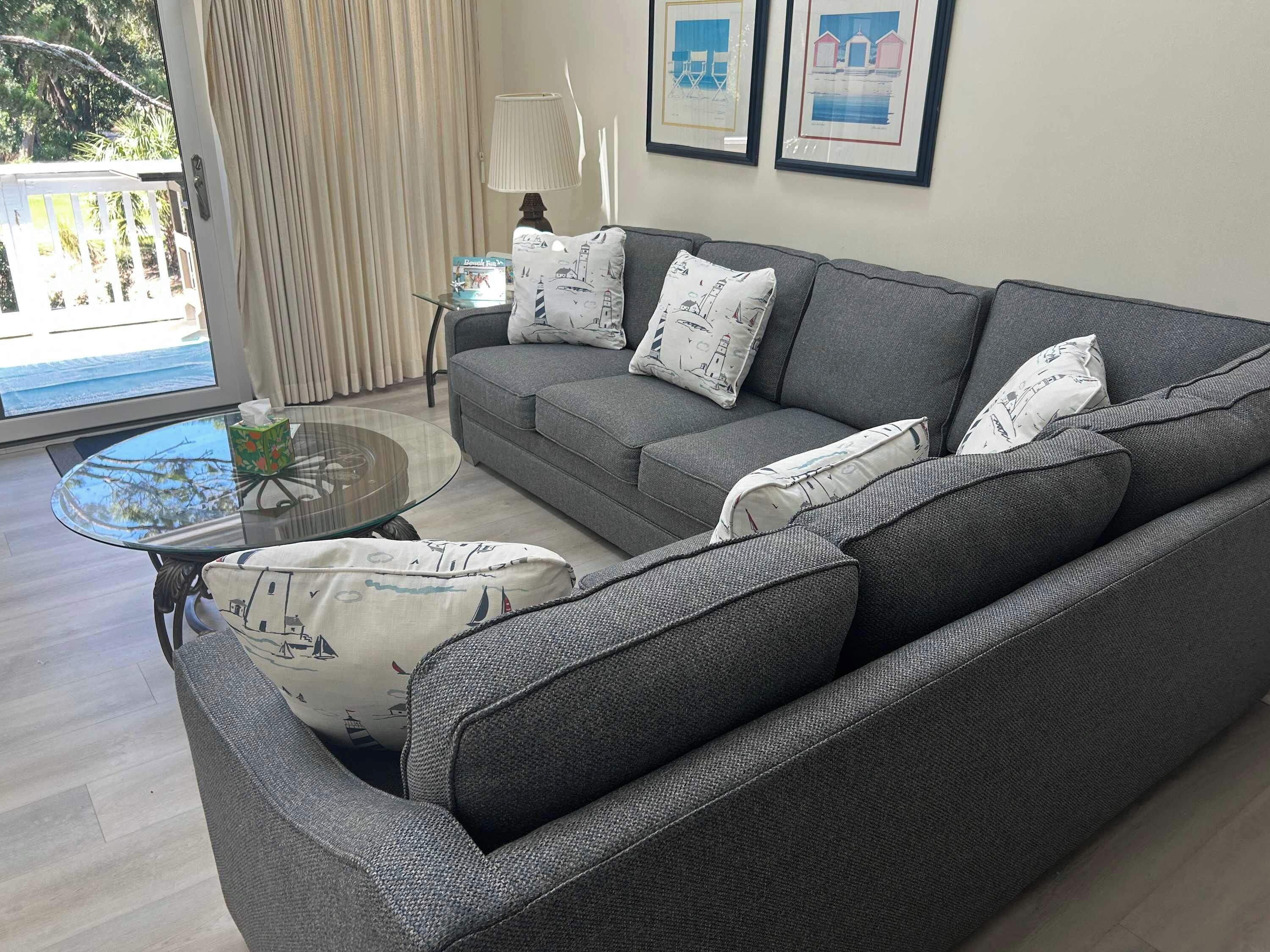Ein modernes Wohnzimmer mit einem grauen Sofa, gemusterten Kissen, einem Glastisch und gerahmten Kunstwerken an der Wand.