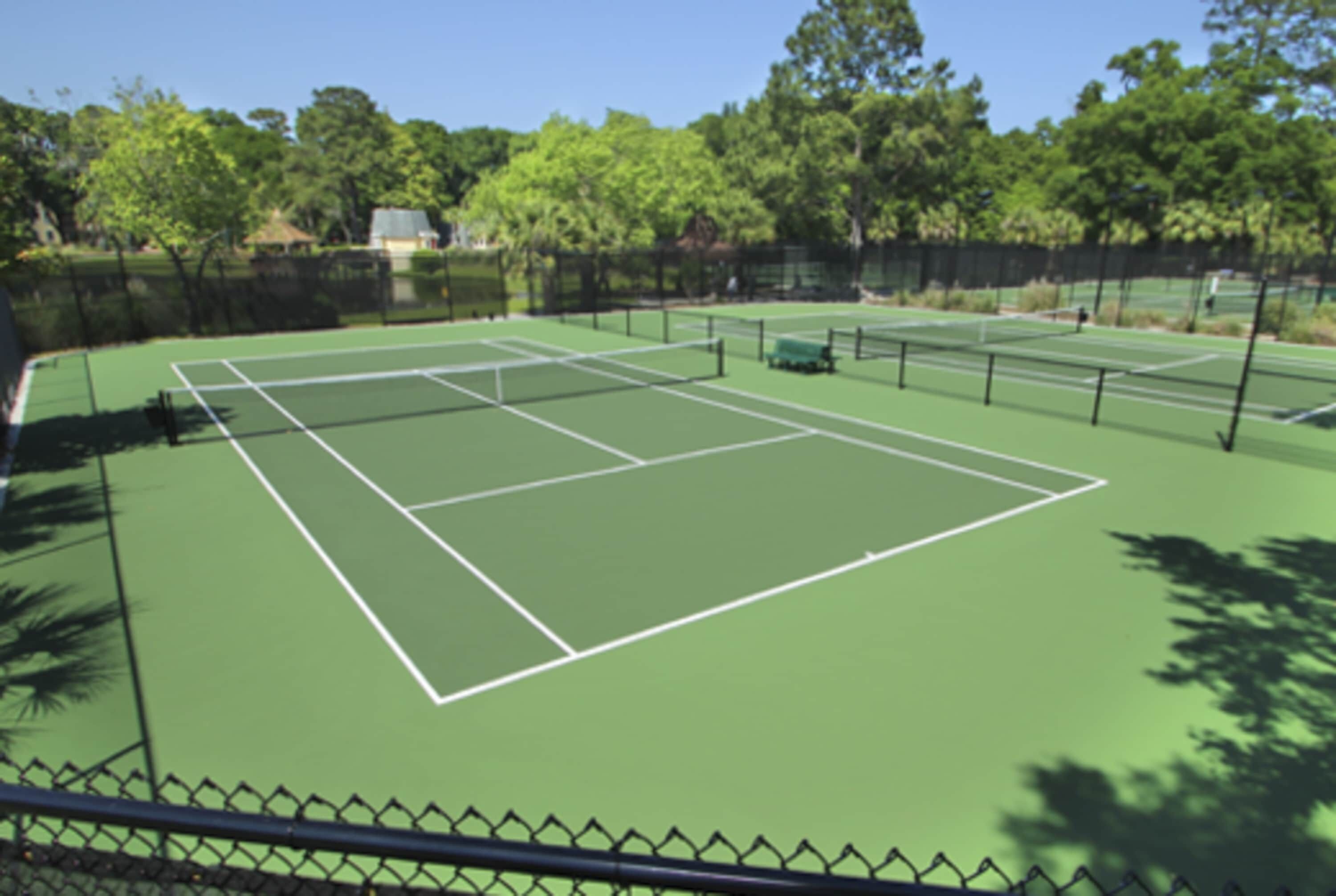 A tennis court with a fence, trees, and a clear sky.