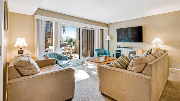 Ein Wohnzimmer mit beigefarbenen Sofas, einem Couchtisch, einem Fernseher und einer Schiebeglastür, die zu einem Außenbereich führt.
