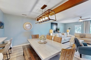 A dining area with a long table, wicker chairs, a chandelier, and a clock on the wall. - Stylish Home with Private Deck, Shared Pool, and access to Dining and Shopping (Hilton Head)