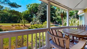 A covered outdoor area with a wooden table and chairs, surrounded by a white railing, overlooking a landscaped garden with various trees and shrubs. - Island Bliss Near the Beach with Golf On Site, Shared Pool,Hot Tub & Private Deck (Hilton Head)