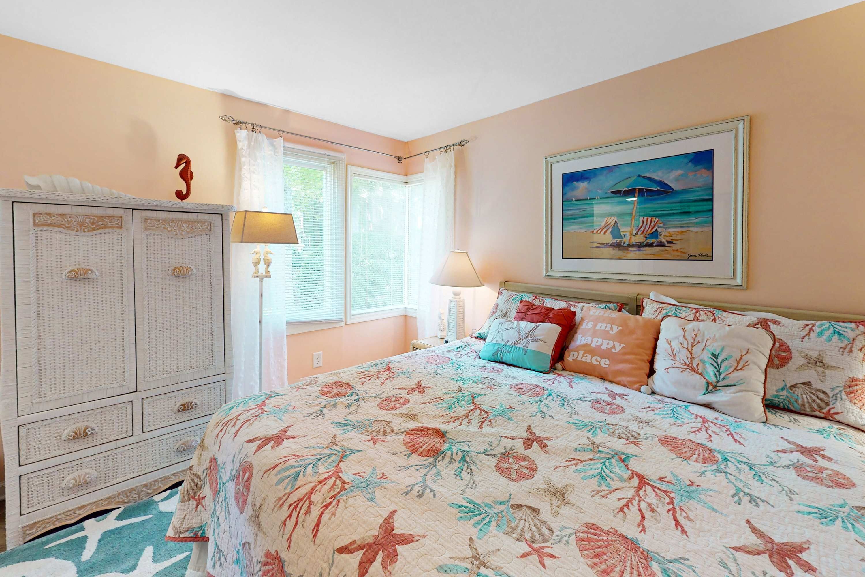 Une chambre à coucher avec une couvre-lit à motifs floraux, un tableau représentant une scène de plage et une commode avec des tiroirs.