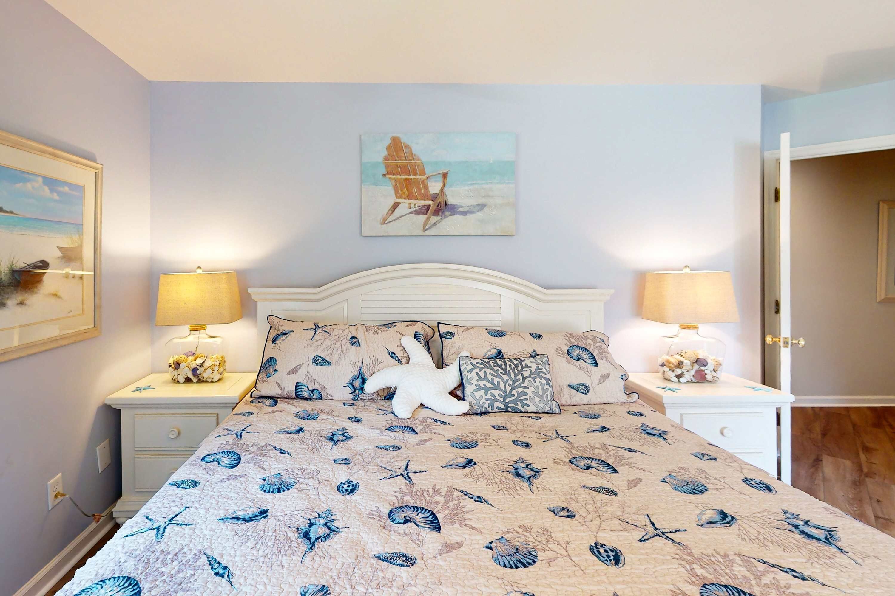 Une chambre à coucher avec un lit, deux tables de chevet, une lampe sur chacune, un tableau au mur et une porte sur le côté droit.