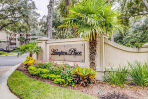 A sign reading 'Hampton Place' with a landscaped garden featuring various plants and a palm tree. - Corner condo with panoramic view & wraparound deck, just steps from the water (Hilton Head)