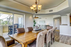 A dining area with a wooden table, chairs, a chandelier, and a view through large windows. - Corner condo with panoramic view & wraparound deck, just steps from the water (Hilton Head)