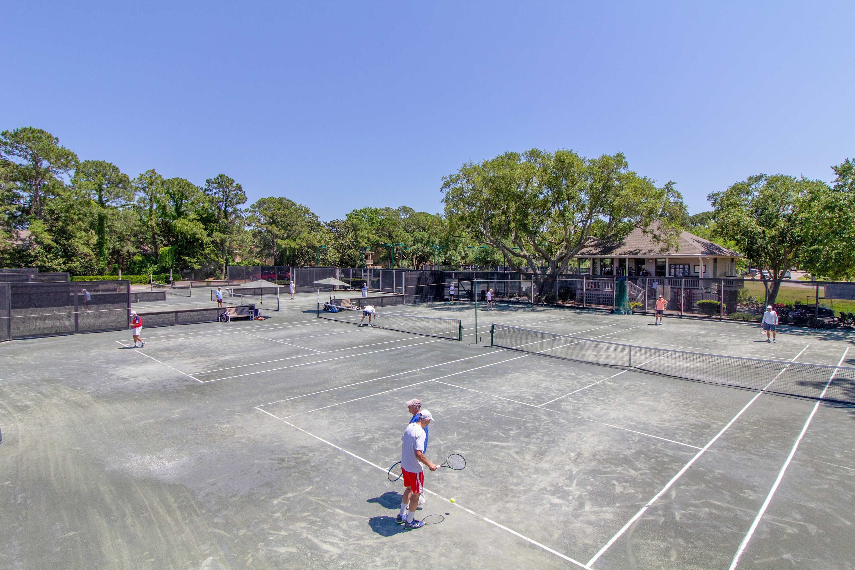 Un court de tennis où jouent plusieurs personnes, une zone couverte pour les spectateurs et des arbres en arrière-plan.