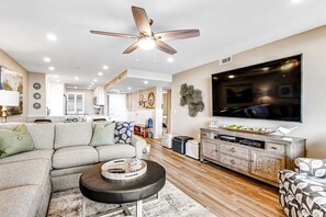 A modern living room with a sectional sofa, a coffee table, a TV, and a ceiling fan. - Cozy oceanfront villa, tennis courts, hot tub, shared pool and ocean view (Hilton Head)