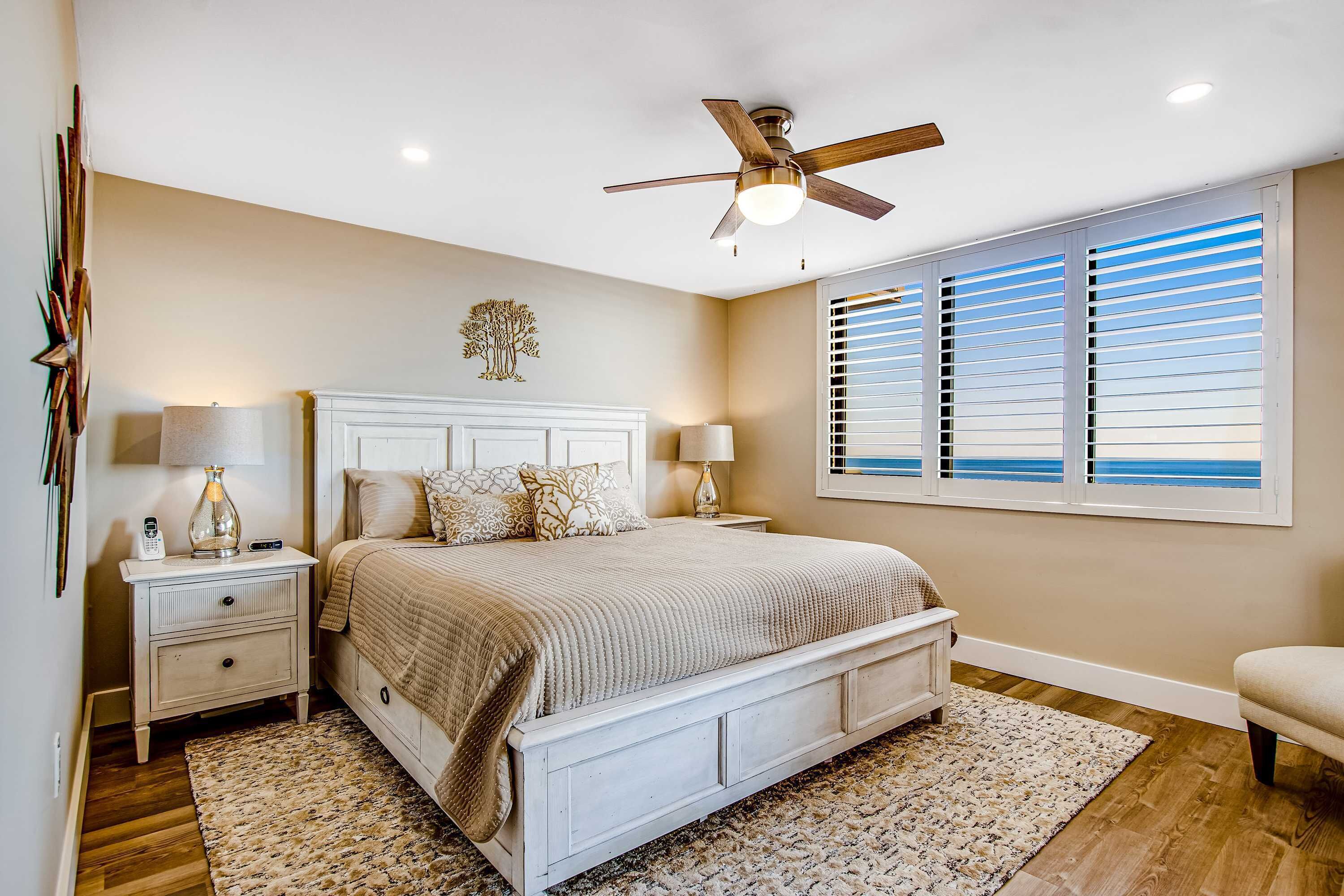 A bedroom with a bed, nightstands, a ceiling fan, a window with blinds, and a decorative wall piece.