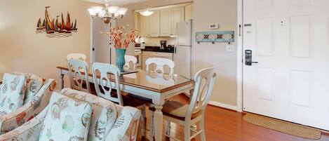 A dining area with a glass table, white chairs, a vase with flowers, and a kitchen in the background.