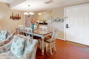 A dining area with a glass table, white chairs, a vase with flowers, and a kitchen in the background. - Second-floor condo with balcony, shared outdoor pool, central AC (Hilton Head)