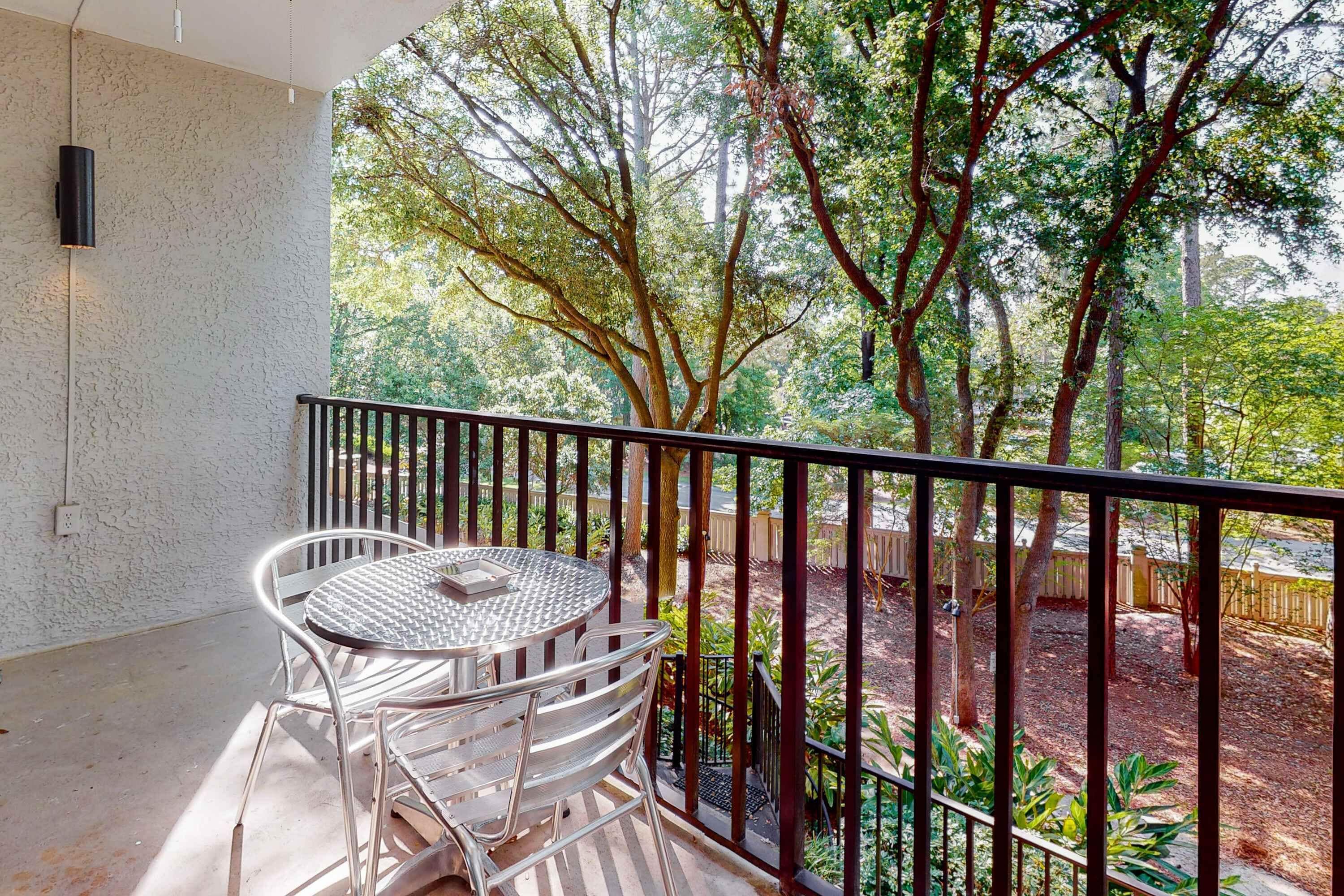 A balcony with a round table and chairs, a black railing, and a view of trees and a garden.