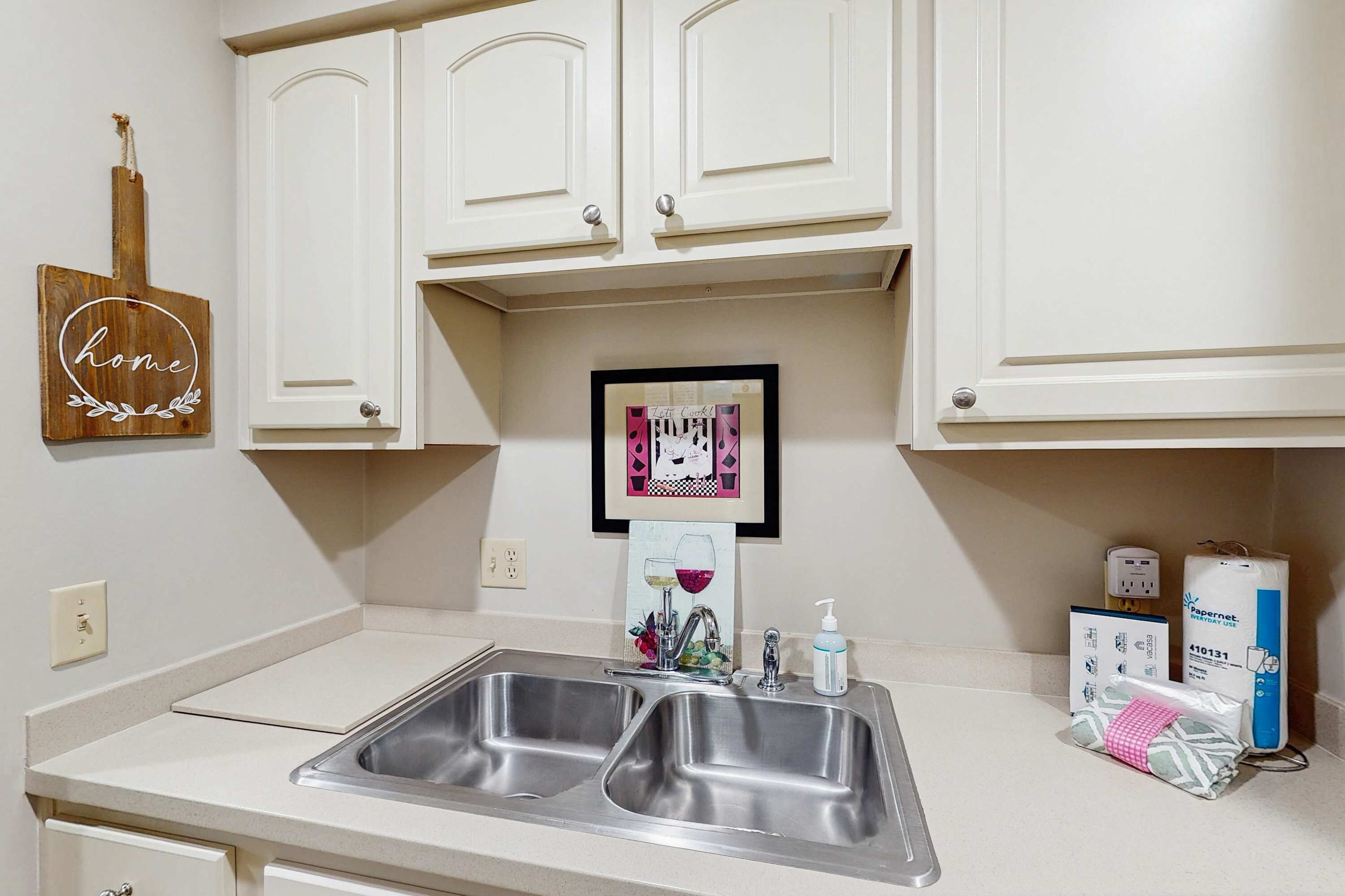 A kitchen with two stainless steel sinks, white cabinets, a framed picture, and a small table with a bottle and a bag.