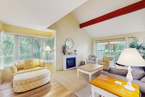 A living room with a sofa, a coffee table, a fireplace, and large windows. - Villa with saltwater pool, golf, firepit, hot tub, & private beach access (Hilton Head)