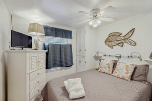 A bedroom with a bed, a TV, a ceiling fan, a wall decoration, and a window with curtains. - Beach condo with Ocean Views, outdoor pool & balcony - Walk to Beach (Hilton Head)