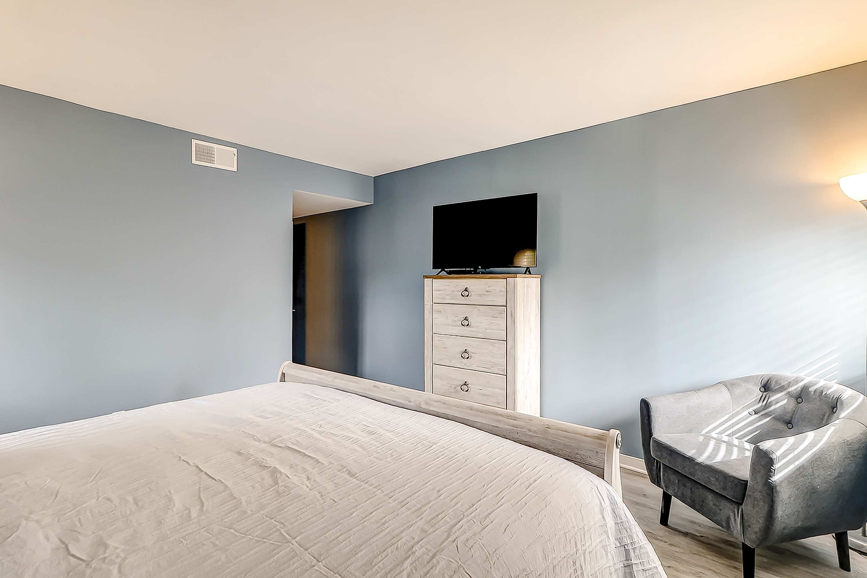 Un dormitorio con una cama, un tocador, una silla y un televisor en la pared.