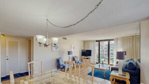 Ein modernes Wohnzimmer mit einem Glastisch, blauen Stühlen mit Muster, einem blauen Sofa, einem Fernseher und einem großen Fenster mit Vorhängen.