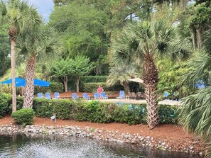 A pool area with lounge chairs, palm trees, and a water feature. - Cozy condo with tennis, pool, beach access, & private furnished balcony (Hilton Head)