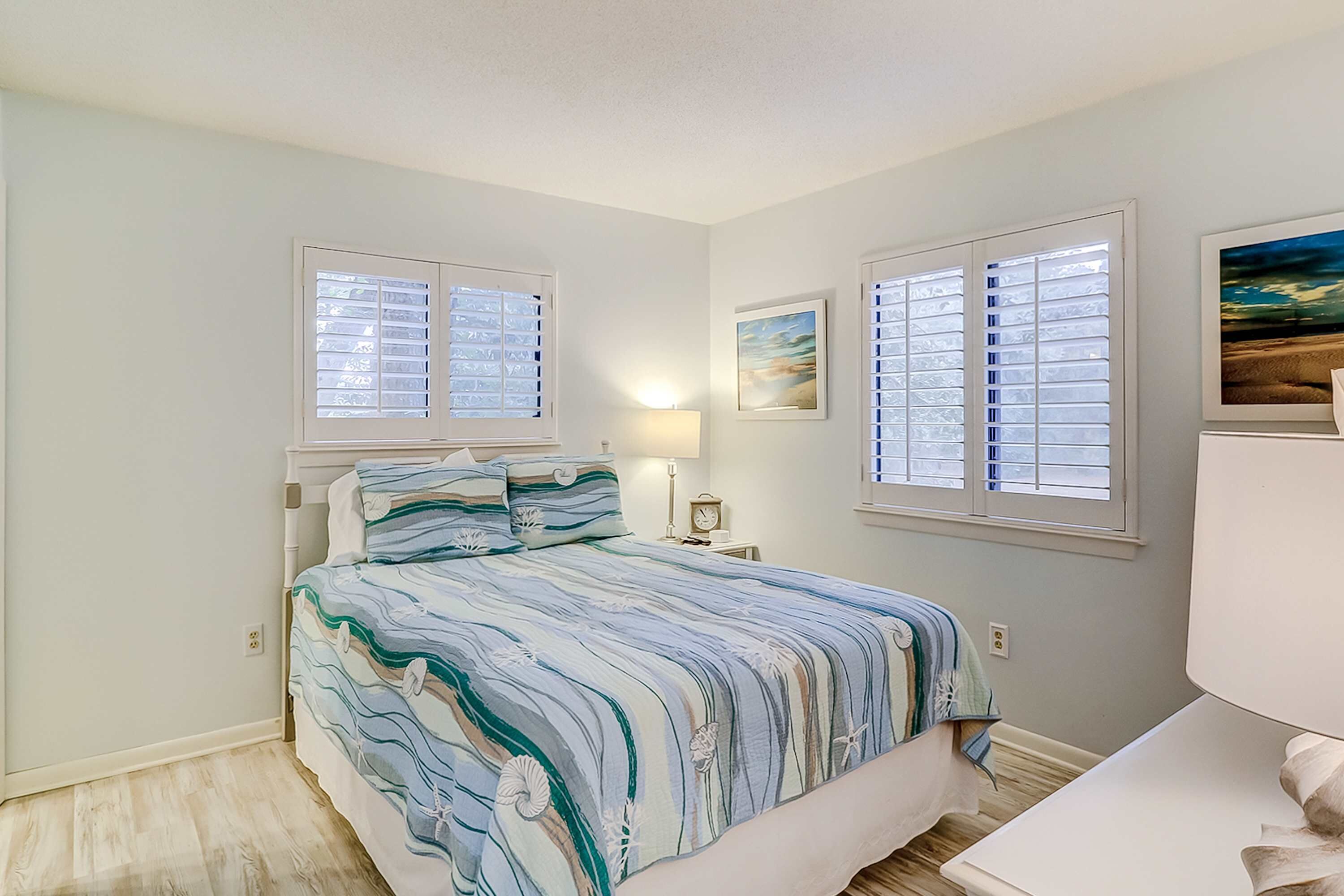A bedroom with a bed, two windows with blinds, a nightstand, a lamp, and a framed picture on the wall.
