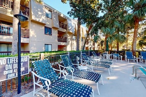A poolside area with lounge chairs, a sign, and a building with balconies. - Charming third-floor condo- private balcony, shared pool, and steps to Coligny Plaza and the beach (Hilton Head)