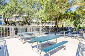 Ein Außenbereich mit einer Terrasse, Picknicktischen und Bänken, umgeben von einem Metallzaun, mit Bäumen und einem Gebäude im Hintergrund.