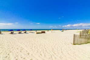 Une plage de sable fin, avec des chaises longues et des parasols, une clôture en bois et un ciel bleu azur.