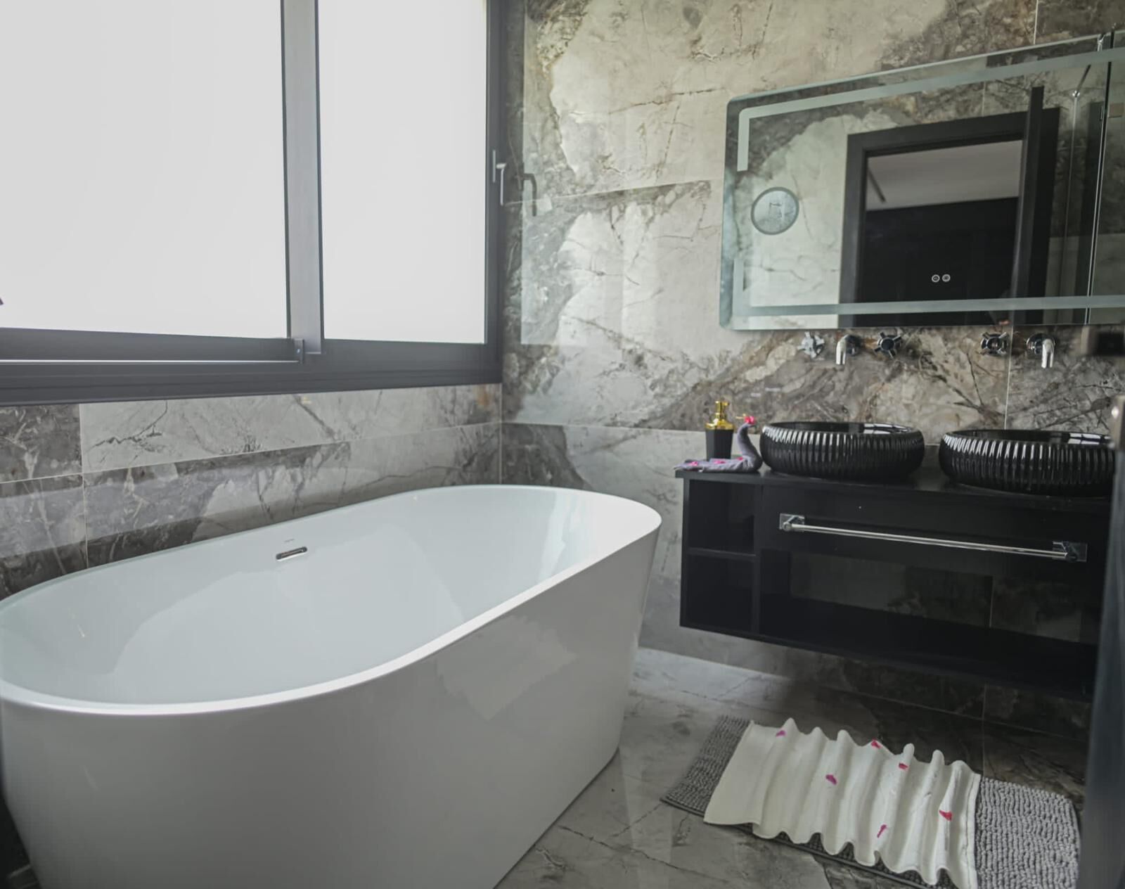 Une salle de bain moderne avec une baignoire sur pieds, un meuble-lavabo en verre avec deux vasques en céramique noire et un mur recouvert de carreaux de marbre.