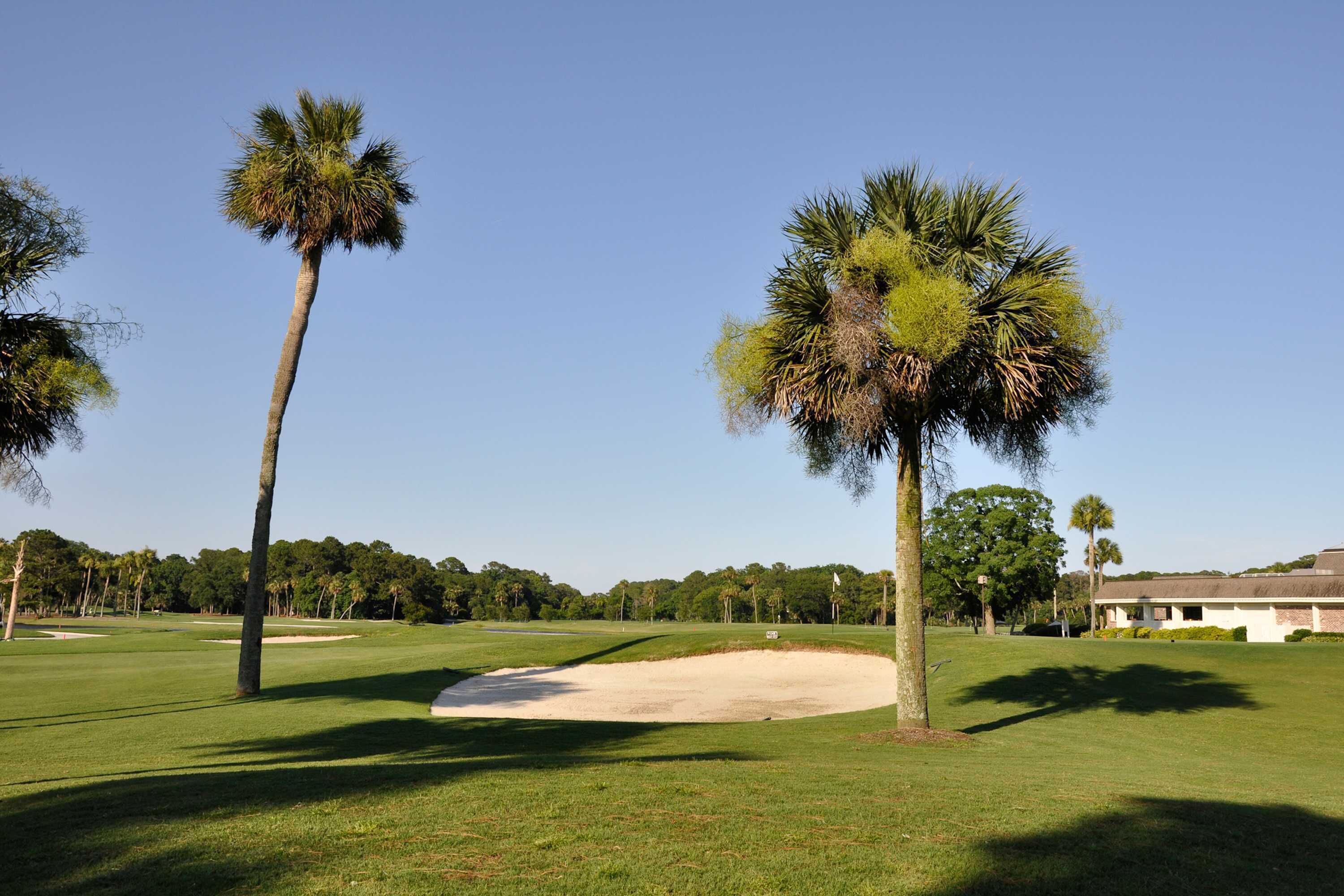 Een golfbaan met zandbunkers, palmbomen en een clubhuis op de achtergrond.