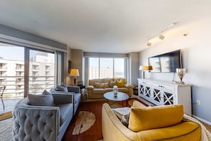 A modern living room with a sofa, armchair, and coffee table, a flat-screen TV, and a large window with a view of buildings. - Family friendly home with an ocean view plus shared pool & hot tub, near the beach (Hilton Head)