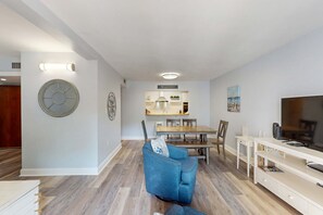 Ein modernes Wohnzimmer mit blauem Sessel, einem Essbereich mit Holztisch und -stühlen sowie einem Fernseher auf einem weißen Schrank.