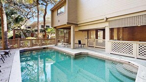 Ein Pool mit Holzterrasse, einer überdachten Sitzgelegenheit und einem Gebäude mit heller Fassade.