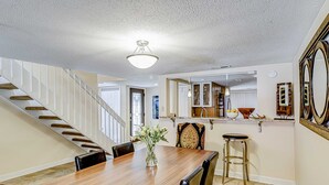 Un comedor con mesa de madera, sillas, un florero con flores, una escalera y una zona de cocina con armarios y un espejo.