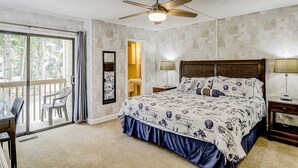 Un dormitorio con cama, ventilador de techo, balcón con sillas y vista a árboles.