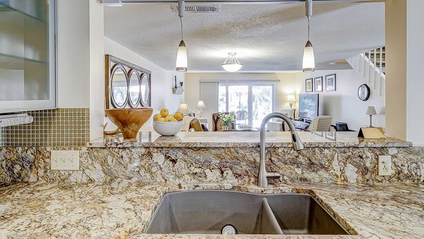 Una cocina con encimera de granito, pileta de acero inoxidable, iluminación colgante y vista al área de estar.
